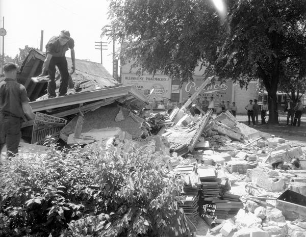 A worker is sorting through the rubble from the collapsed Kleinheinz Pennco service station (Pennsylvania Oil Company), 702 S. Park Street.  People in the background are looking on from under the shade of a tree, with Kleinheinz Pharmacies and Pennco billboards in the background.