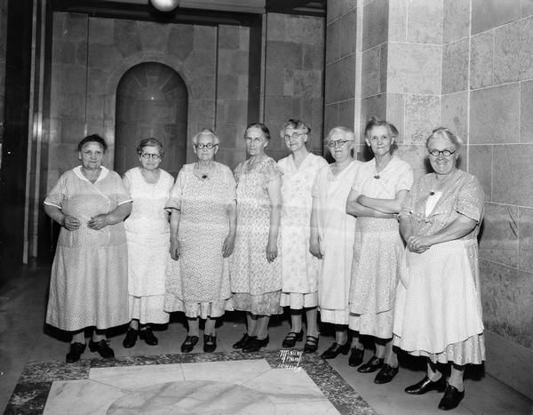 Group portrait of eight Wisconsin State Capitol scrubwomen. Left to right: Mrs. Mary DeRenzo, Mrs. Elizabeth Weddig, Miss Mary McCloskey, Mrs. Ingeborg Sidwell, Mrs. Margaret Harbort, Mrs. Olivia Kver, Mrs. Katherine McKune, Mrs. Catherine Conlin. Mrs. DeRenzo is the oldest scrubwoman.