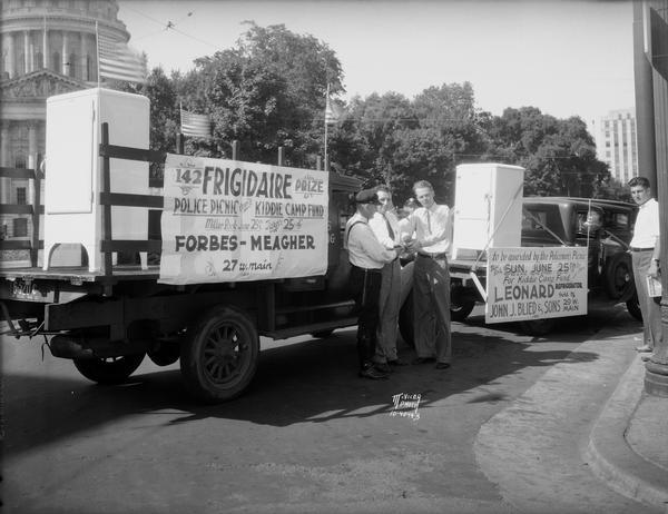 Police officer selling raffle tickets to two men for refrigerators displayed on truck beds. One sign reads "Frigidaire prize. Police picnic benefit for Kiddie Camp fund, Miller Park, June 25, tickets 25 cents at Forbes Meagher. 27 W. Main St." The other sign reads "To be awarded by the policemen's picnic for Kiddie Camp Fund. Leonard Refrigerator sold by John J. Blied & Sons, 28 West Main Street."