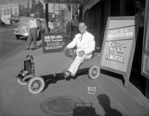Harold Knudsen sitting on the National Junior Racer outside the Capitol Theatre, the prize for a contest conducted by the Fiore Coal and Oil Co., Phillips "66" gasoline stations, and the Capitol Theatre. The promotional sign reads: "Look! Boys and Girls. Free! This real automobile, Pep-Zip-Go-and how! Runs 60 miles on a gallon of Phillips 66 gas. Powerful 4 cycle motor, two speeds forward and reverse, 5 wire loads and real balloon tires."