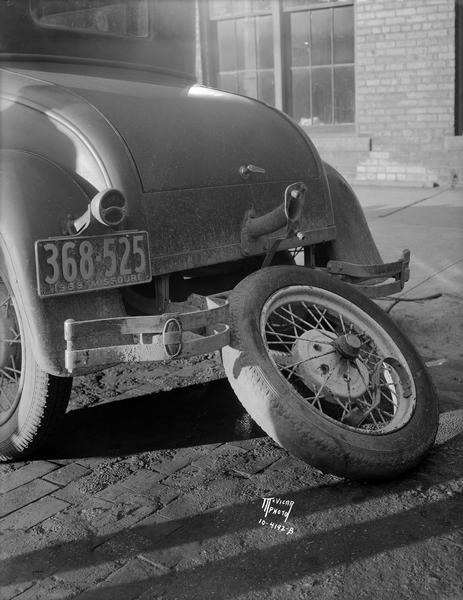 View of rear end damaged Ford Coupe, with the spare tire broken off and leaning against the car.