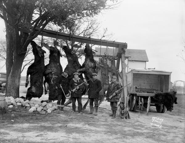 Four men posing with their dog and hunting trophies — moose, deer, bear, and fish hanging in the farmyard.