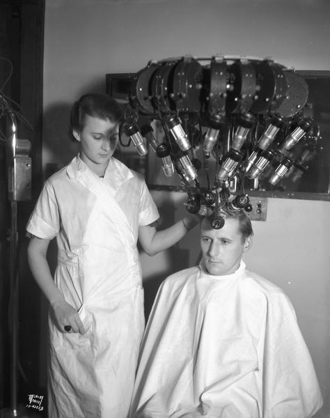 Cedric Parker, Capital Times reporter, gets his hair curled with a permanent wave machine attached to his hair, at Mrs. Wengel's Marinello Shop, 125 State Street. Lucille Bump, beauty operator, is standing by.