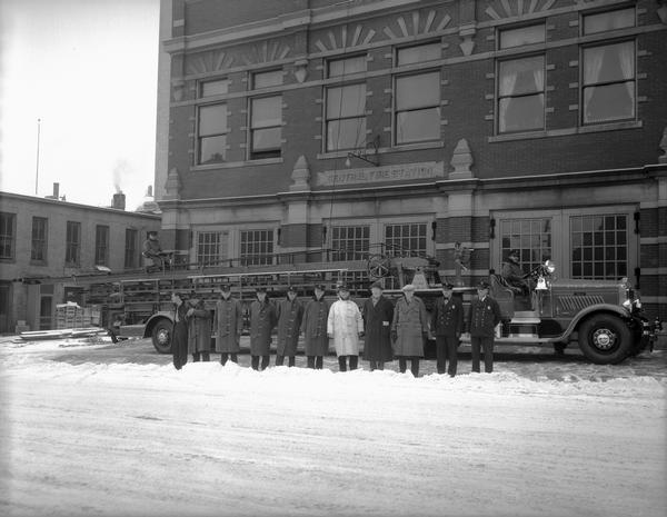 Madison Fire Department's new gray $16,200 aerial fire truck parked in front of the Central Fire Station, 18-20 South Webster Street. Roy Hurling is driving and Jim Williams is acting as tiller man, with eleven fire fighters lined up in front of the truck in the snow.