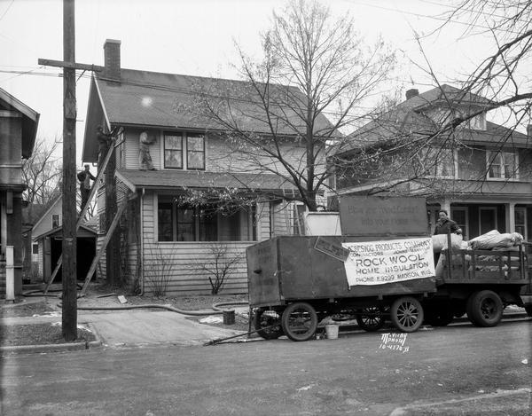 A Johns-Manville Contractors Asbestos Products Company Rock Wool home insulation truck and trailer is parked in front of a house, with workers on ladders and the roof, blowing rock wool insulation into the house.