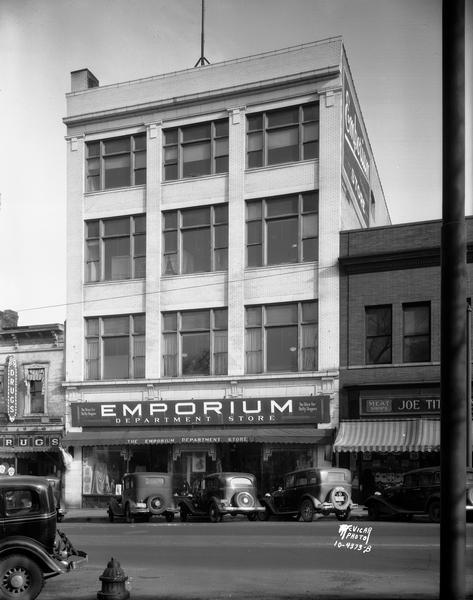 Exterior of the Emporium Department Store at 15-17 North Pinckney Street, also showing Joe Tittle Meat Market, Rennebohm's drug store, and automobiles angle parked along the street.