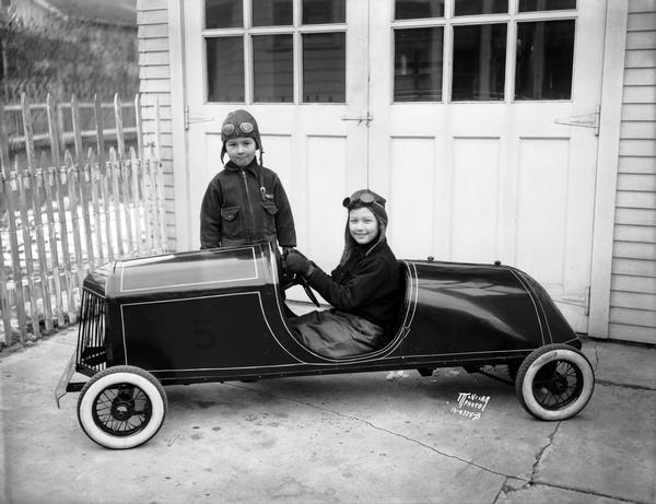A small race car built by Samuel Olson at the Wisconsin Oil Burner factory, 1134 Regent Street is enjoyed by his sons, Donald Olson, seated, and Gordon at their home at 2221 Fox Avenue. The boys are wearing aviation-style helmets and goggles.