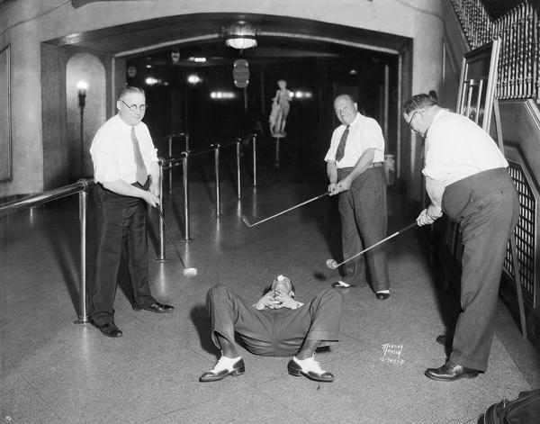 Men posing during a golf stunt, as they try to hit a golf ball off a man's nose while he is lying on his back on the floor in the lobby of the Orpheum Theatre. This was a promotion for the Fanchon & Marco show.