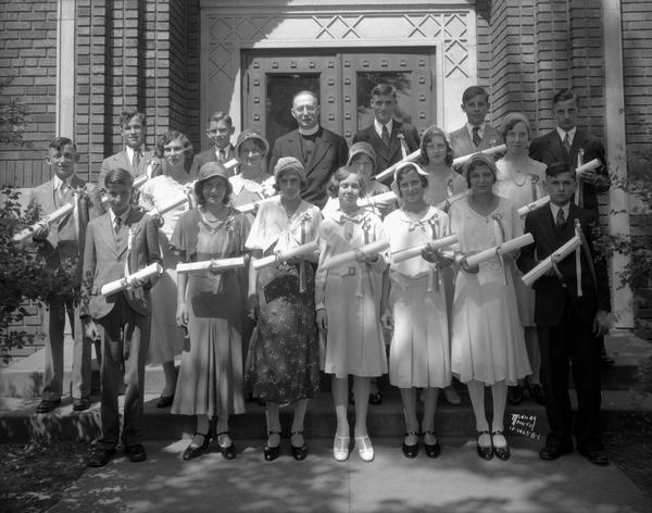 St. Aloysius school with children and their diplomas, and a priest in front of the school door.