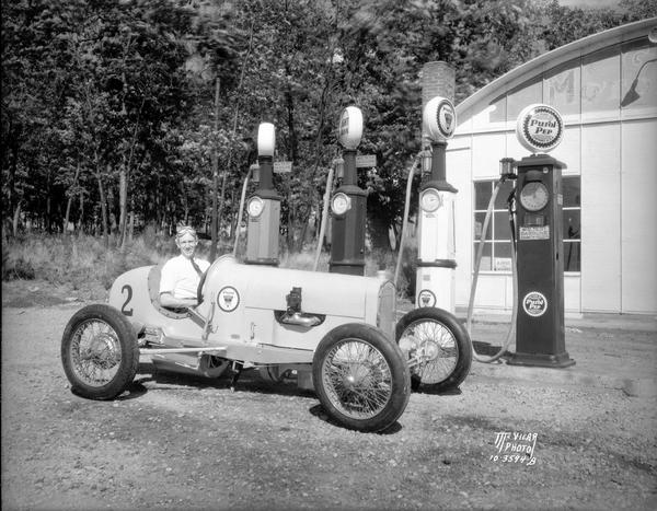 Giles Hanson seated in his racing car in front of a gas station with Purol Pep gas pumps.