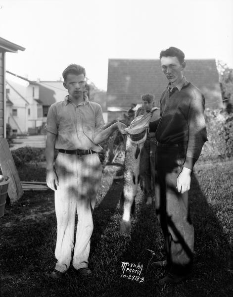 Two men in a backyard are holding up a large pickerel fish that they presumably caught.
