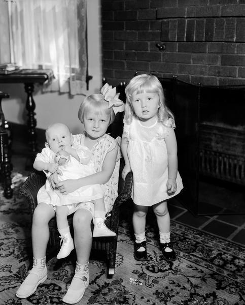 Portrait of Lois Kerl's three children: one daughter is sitting and holding a baby, and the second daughter is standing on the right in front of the fireplace.