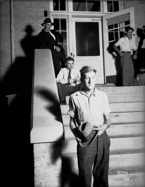 Alfred Deering, on the steps in front of the Doylestown Bank, holding his hat. He received a wound to the head during the Doylestown bank robbery. Men are standing and sitting behind him at the top of the steps near the entrance to the bank.