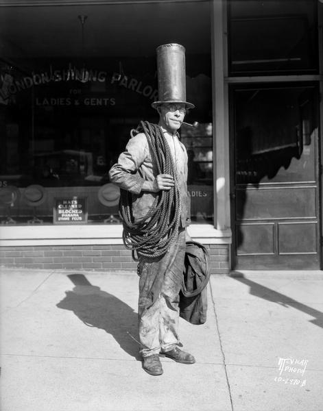 Portrait of George Stahl, chimney sweep, with a pipe in his mouth, and wearing a tall hat and carrying a bag and a rope around his shoulder. He is standing in front of the London Hat Cleaners, 230 King Street.