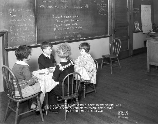 Four deaf children are sitting at a table playing at life experiences at Doty School. Sentences on the blackboard describe their activity to help them learn language. Caption reads: "Deaf children play actual life experiences and then are given language to talk about them. Madison Public Schools."