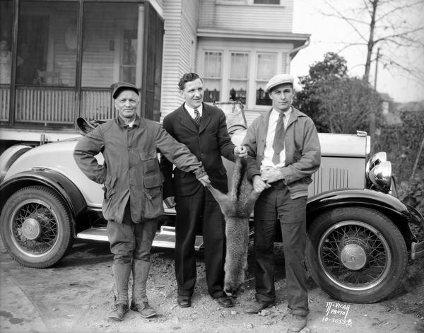 Three men (including Hilgers) proudly display their trophy fox. An automobile is behind them, and a house is in the background.