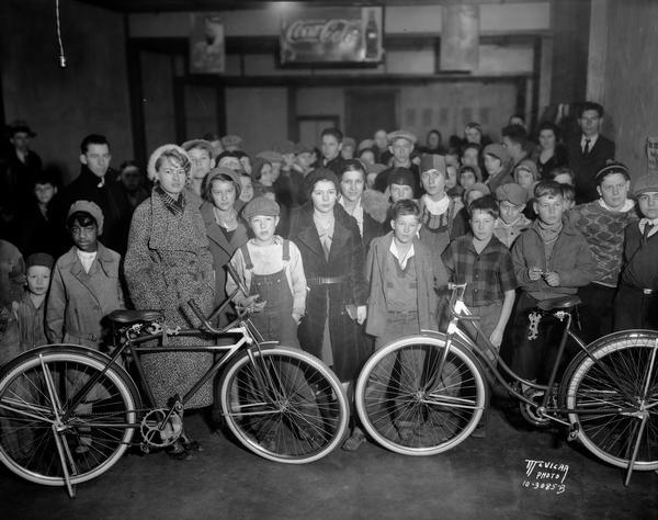 A group of children and adults observe the awarding of bicycles as prizes in the Madison Label Saving contest.