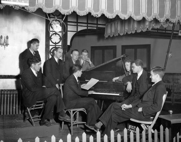 Group portrait of nine orchestra members gathered around a grand piano at the Studio ballroom, 237 West Gilman Street.