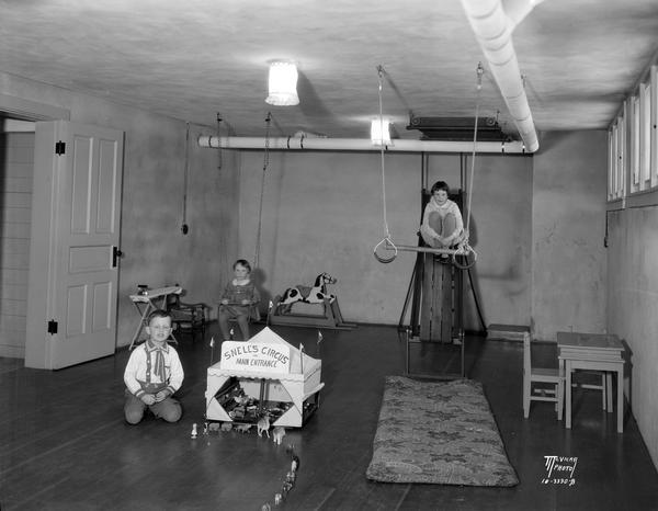 View of the basement playroom at Chester and Louise Snell's home, 822 Miami Pass. The three children are playing with Snell's miniature circus, rocking horse, slide, swing, and trapeze.