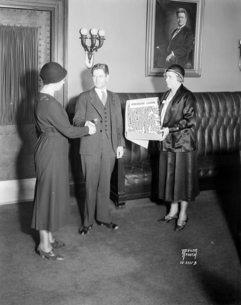 Governor Philip F. La Follette buying a "buddy poppy" from two women in behalf of the American Legion.  The governor is standing in his Wisconsin State Capitol office near a portrait of his father, Robert M. La Follette, Sr.