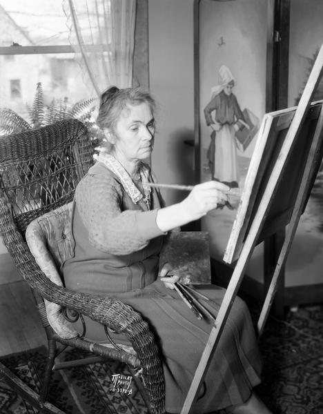 Artist Frances Hawkins sitting in a wicker chair and painting at the easel in her studio.