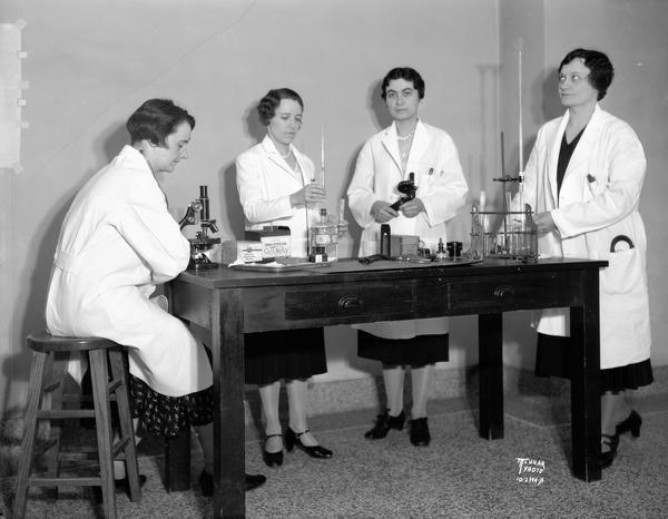 A group of four women medical students wearing laboratory coats at Wisconsin General hospital.  Left to right, Mrs. A.F. Kyhos is sitting at a microscope with a stethoscope around her neck, May A. Davies is holding a pipette and test tube, Alice D. Watts is holding a hemoglobin meter, and Addie M. Schwittay is holding a burette.