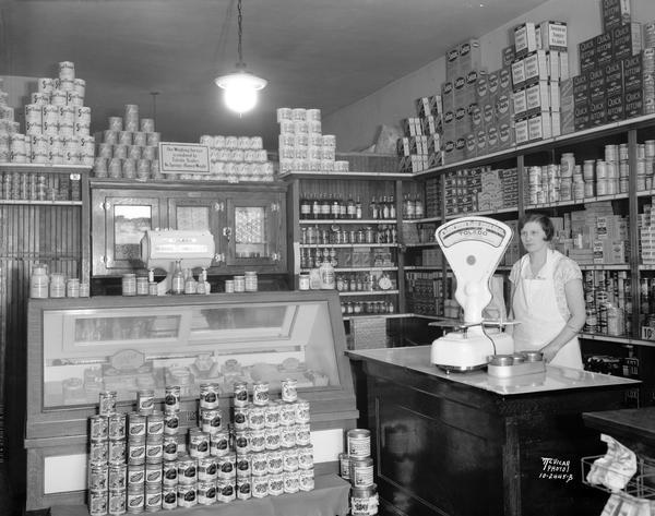 Interior of the Avenue Food Shop, located at 2427 University Avenue, with stacks of canned goods and a woman employee using a Toledo scale.