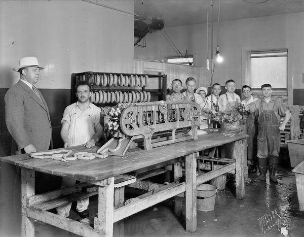 Sausages coming off of the sausage machine at the Madison Packing Company, 307 West Johnson Street. Standing around the machine are eight workers and Oswald B. Neesvig.
