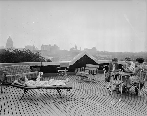 Three University of Wisconsin-Madison coeds are playing cards, and one woman is reading, on the roof of Ann Emery Hall, 265 Langdon Street, an independent women's dormitory named for the first Dean of Women (1897-1900) Ann Emery Allinson (1871-1932). The Wisconsin State Capitol and Madison skyline are in the distance.