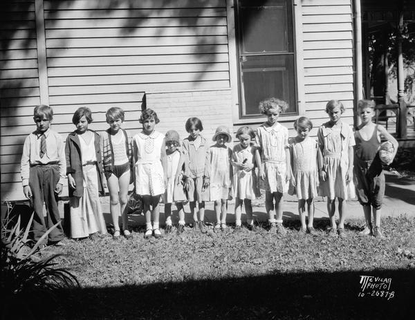 Twelve children dressed in clothing ranging from bathing suits to a necktie posing as they wait in a yard to leave for Kiddie Camp.