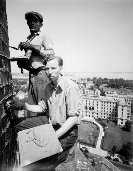 Steeplejacks Don Coffey, in the foreground, and Bruce Russell, in the cap, repointing the University of Wisconsin heating plant chimney. The photograph was made from a scaffold suspended 192 feet in the air. In the background, the elevated view includes the University of Wisconsin Hospital, Elizabeth Waters women's residence hall, the Home Economics building, Lake Mendota, and Picnic Point.
