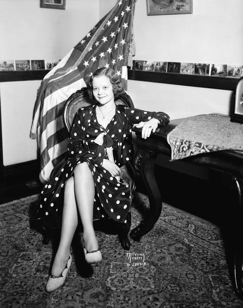 Disabled American Veterans Ball queen contestant wearing a polka dot dress. She is sitting in a chair in front of an American flag.