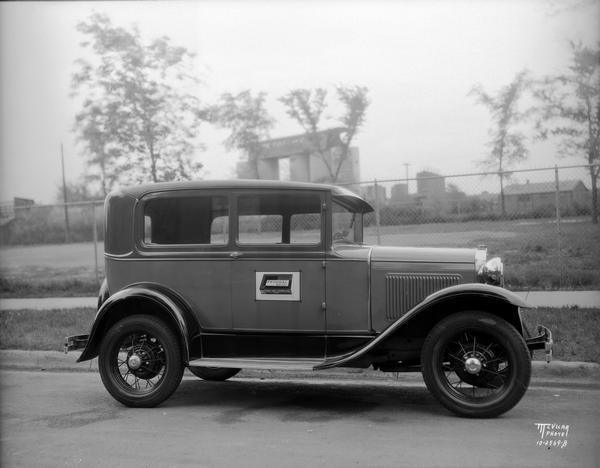 An automobile advertises Federal Match Company. There is a sign on the passenger side door that reads: The sign reads: "Federal Matches, Federal Match Corporation, Chicago." There are storage tanks in the distance.