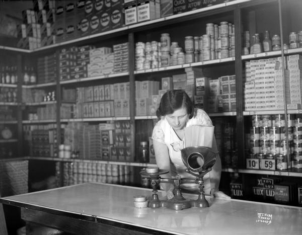Interior of the Avenue Food Shop, 2427 University Avenue, with a woman weighing items on an old balance beam scale.