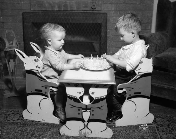 Richard and Malcolm McVicar celebrating Richard's fourth birthday while sitting in front of a fireplace with a birthday cake and candles. They are sitting at a child-sized table and chairs with a rabbit motif.