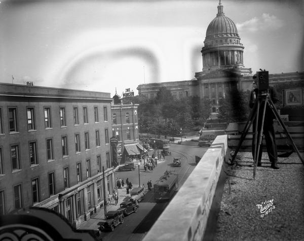 The corner of State Street and Capitol Square from the roof of the Leath Furniture Store, with a view of the 100 block of State Street, the Wisconsin State Capitol, Commercial State Bank, and the Wisconsin Life Insurance Company building. Mr. Knutson is standing on the roof behind a movie camera on a tripod.