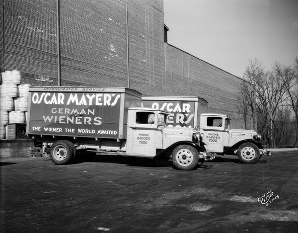Two Oscar Mayer delivery trucks made by Diamond T waiting at a loading dock. One of the trucks has a sign for "German Wieners." There is a stack of barrels on the loading dock.