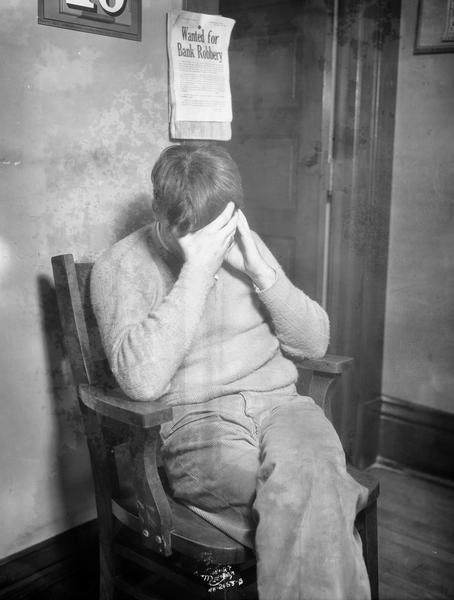 Myron Stephanson, accused burglar, holding his head in his hands while sitting under a notice titled: "Wanted for Bank Robbery" at the police station.
