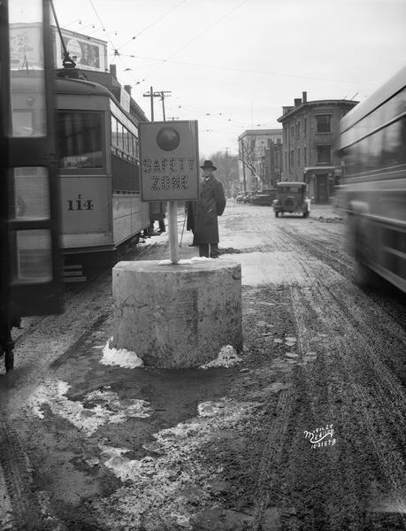 A man stands at a safety zone in the middle of West Mifflin Street between lanes of traffic at the corner of State Street and N. Carroll Street, waiting to board a streetcar.
