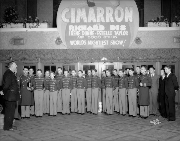 Orpheum and Capitol theatre ushers listening to a talk about fire prevention as they are standing beneath a marquee that advertises the epic "Cimarron."