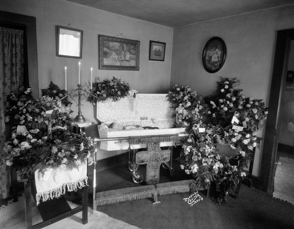 Five-year-old Wilbur Corcoran, son of Frank and Margaret Corcoran, 624 Williamson Street, lying in a coffin surrounded by flower arrangements and a three-tiered candelabra. At the front of the coffin is a religious kneeler.