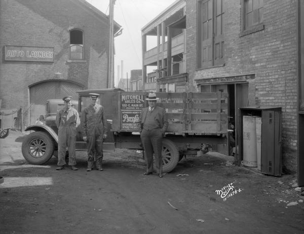 Pyrofax bottled gas agent, Mitchell Sales Company's delivery truck. Two men wearing uniforms and hats are standing near the cab of the truck, and another man wearing a suit and hat is standing near the bed of the truck. They are in an alley at the rear of a store. A sign on a large brick building in the background on the left reads: "Auto Laundry."
