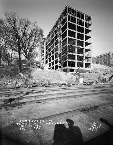 View of the State Office Building, 1 W. Wilson Street, under construction, from Lake Monona. The photographer's shadow and railroad tracks are in the foreground.