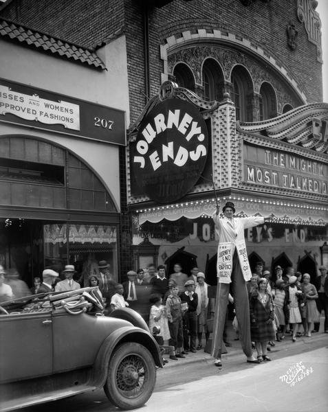 View from street of the facade of the Capitol Theatre at 209 State Street. The marquee is advertising the movie, "Journey's End." There is a crowd on the sidewalk in front of the theatre, and a man on stilts is standing in the street near an automobile.