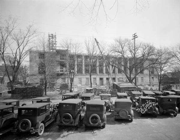 Elevated view looking northeast from Monona Avenue at the front of the U.S. Post Office and Federal Court Building under construction, with a large number of automobiles and a construction crane in the foreground.