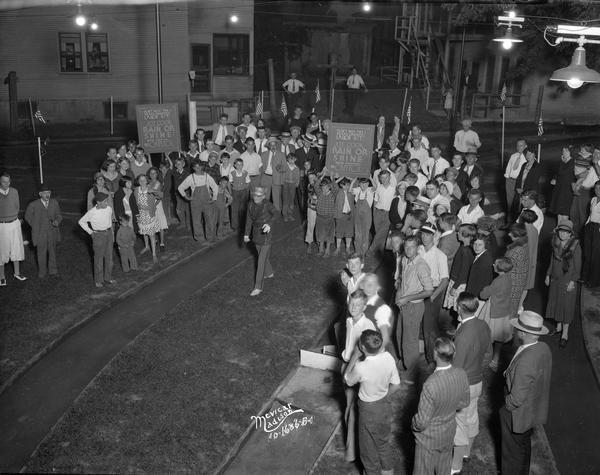 Elevated view of a man in uniform leading a small parade of children who are holding a sign advertising the movie: "Rain or Shine" playing at the RKO Capitol Theatre, while a small crowd watches.