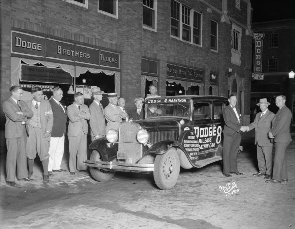 Two men shaking hands in front of Dodge Brothers Trucks and Madison Motor Car Company next to a Dodge 8 Marathon automobile. The mounted spare tire has a sign that reads: "The Greatest Demonstration of Economy-Long Life-Dependability Ever Made by a Motor Car."