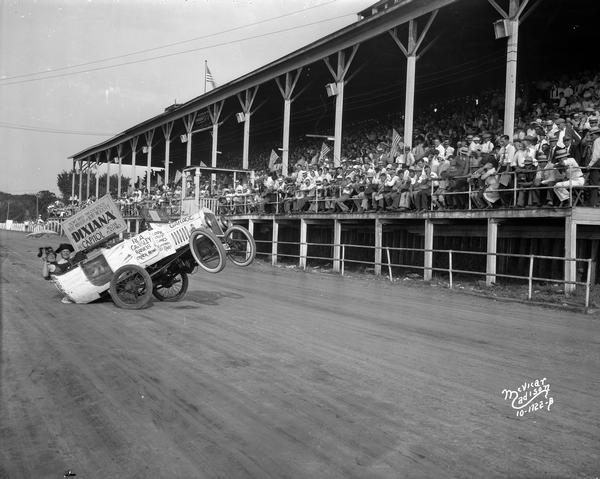 An audience in the grandstand at the Dane County Fair watches two men in a stunt automobile advertising the RKO film, "Dixiana."