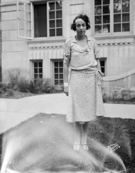 Portrait of Nina Fox standing on a lawn, wearing a dress with a ruffled collar. In the background is a stone building.