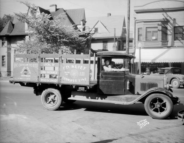 Men are sitting in the cab of the Ed Klief Transfer Line truck, advertising the RKO Orpheum Theatre along with Klief's moving services, in front of the UW Meat Market, 728 University Avenue.